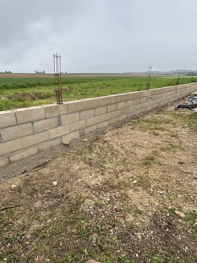 Mur de clôture en parpaings en cours de montage le long d’un champ, armatures verticales des poteaux en attente