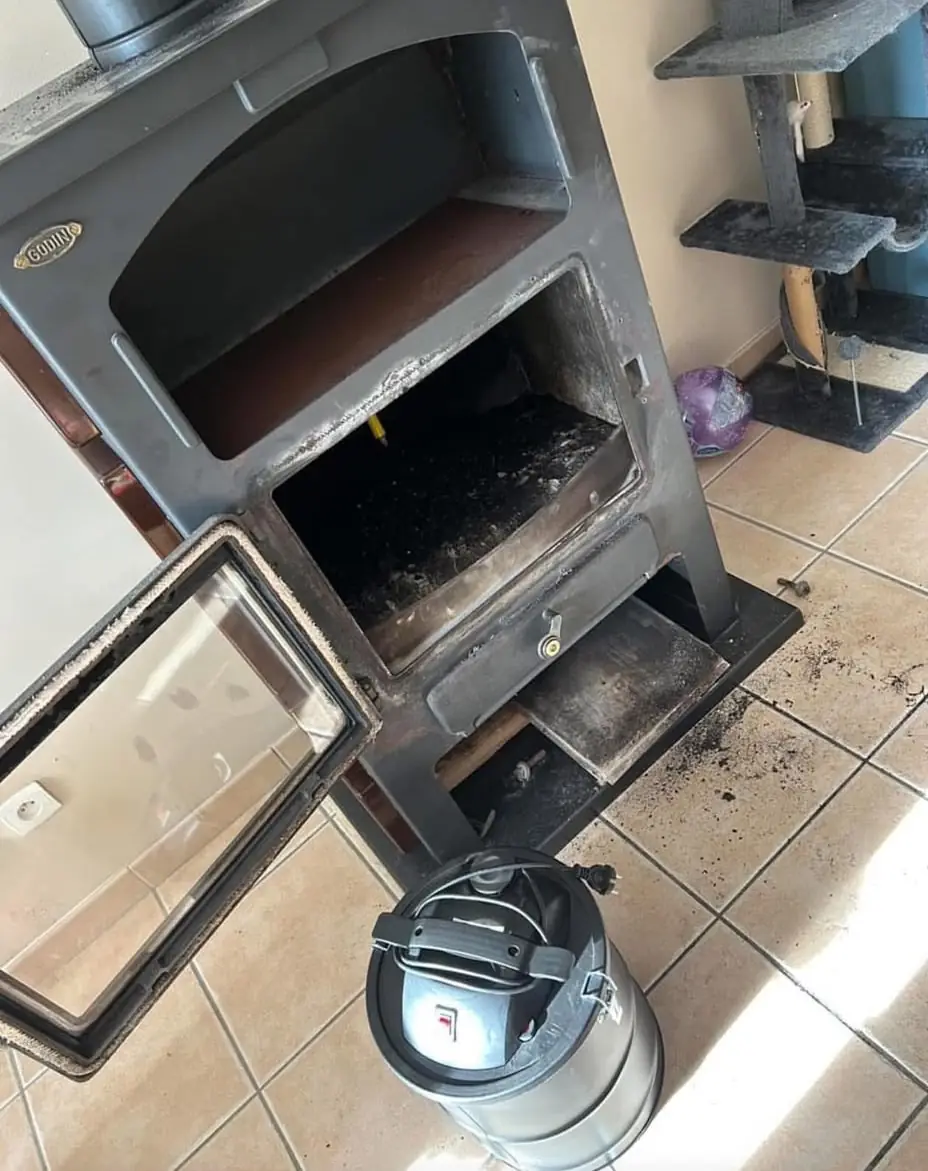 Poêle Godin porte ouverte pendant le ramonage, suie tombée sur le carrelage, aspirateur à cendres au premier plan